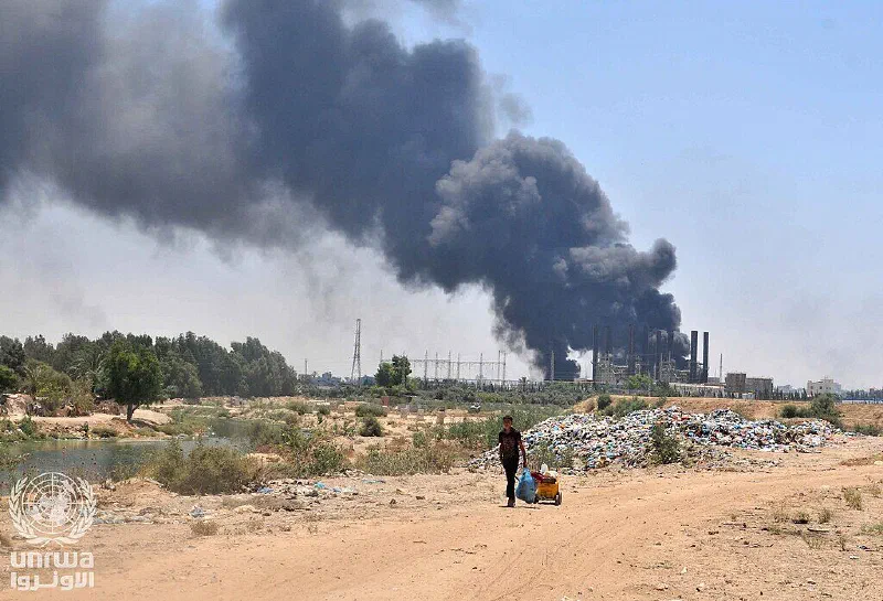 A boy pulls a cart of belongings along a dirt track as smoke rises from Gaza's bombed power plant.