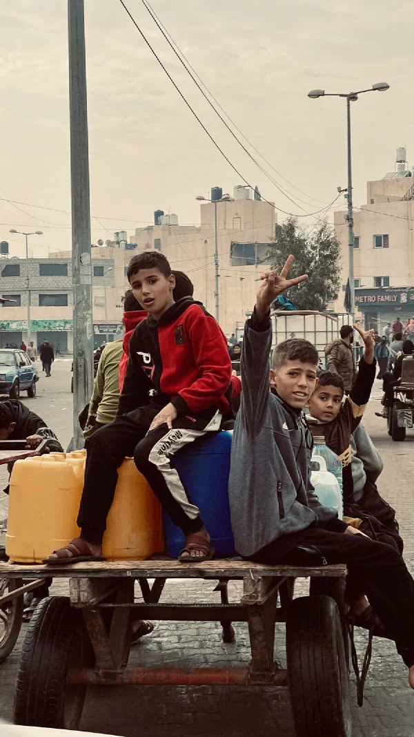 Boys sit with water containers on a cart in a Gaza street, one raising his hand in a peace sign.