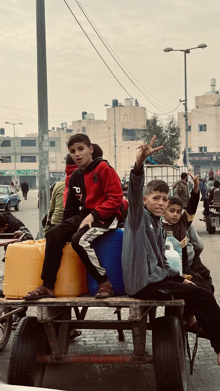 Boys sit with water containers on a cart in a Gaza street, one raising his hand in a peace sign.