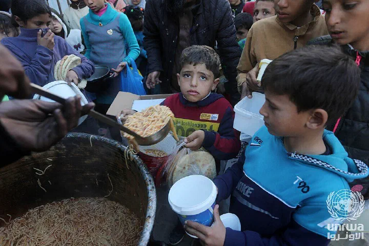 Children hold containers as food is served from a large pot at a community kitchen in Deir el-Balah.