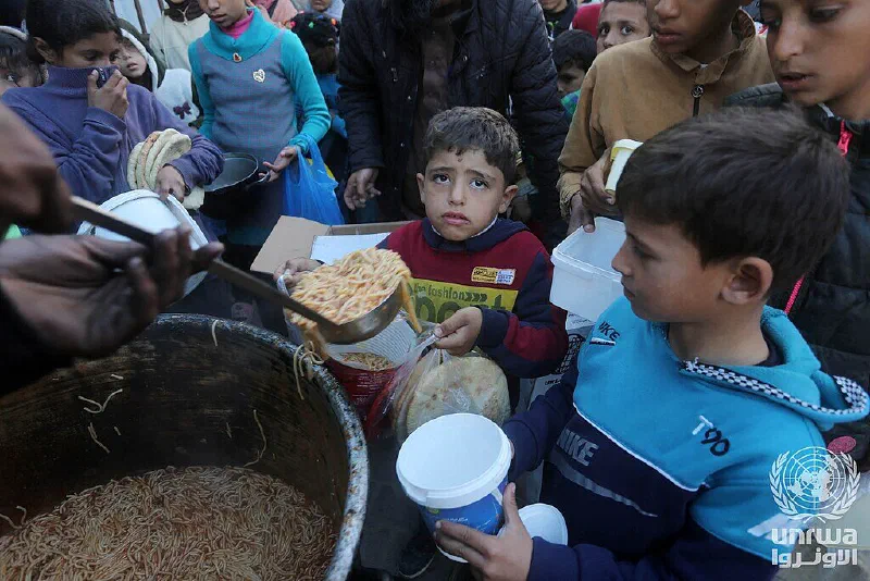 Children hold containers as food is served from a large pot at a community kitchen in Deir el-Balah.